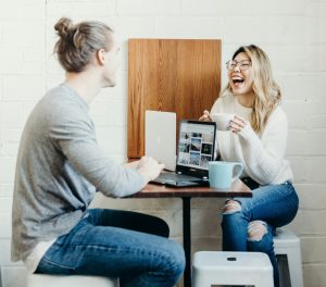 A man and a woman sitting at a table with laptops having a conversation. The woman is laughing